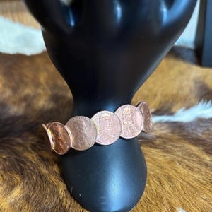 May include: A copper-colored bracelet made of pennies. The bracelet is displayed on a black hand-shaped jewelry holder. The pennies are linked together to form a flexible band. The coins show the profile of Abraham Lincoln.