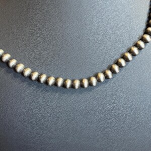 May include: A silver beaded necklace with a matte finish. The beads are round and evenly spaced, creating a simple yet elegant design. The necklace is displayed against a dark background.