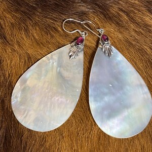 May include: A pair of pear-shaped earrings with a mother-of-pearl finish. Each earring features a small, dark red gemstone and silver-colored metal accents. The earrings hang from silver-colored hooks.