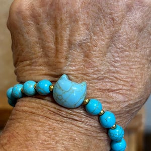 May include: A turquoise beaded bracelet featuring a cat-shaped charm. The bracelet has round turquoise beads separated by small gold-colored beads. The cat charm is light blue with a crackle pattern.