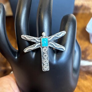 May include: A silver-toned dragonfly ring with a turquoise-colored center stone. The ring features detailed wing designs and spiral accents on the body. The ring is displayed on a black hand.