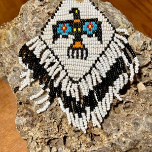 May include: A white and black beaded earring with a beaded bird design and a gold accent. The earring has a fringe of white and black beads.