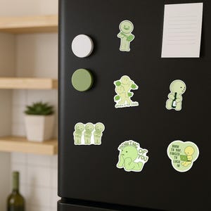 May include: A collection of green stickers with cartoon figures and text on a black refrigerator. The stickers feature various poses and phrases, including "Thinking of You" and "Born to Yap, Forced to Lock In."