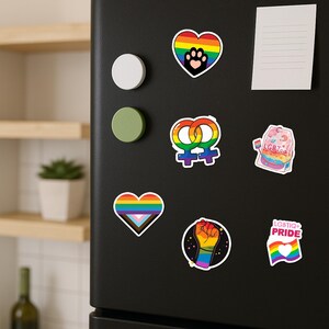 Może przedstawiać: Kolekcja kolorowych naklejek LGBTQ+ pride na czarnej powierzchni. Naklejki zawierają tęczowe serca, odcisk łapy, uniesioną pięść i flagę LGBTQ+ Pride. Inne naklejki przedstawiają symbol żeński i torebkę herbaty z napisem "LGBT Tea".