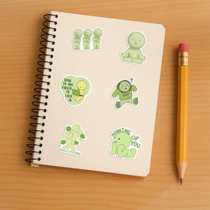 May include: A spiral-bound notebook with several green character stickers and a yellow pencil. The stickers feature various poses and phrases, including "Thinking of You" and "Born to Yap, Forced to Lock In."