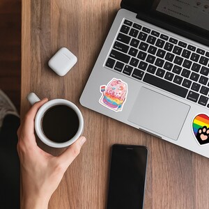 Może przedstawiać: Laptop z naklejką "LGBTea" i tęczowym wzorem. Na drewnianej powierzchni filiżanka kawy, smartfon i bezprzewodowe słuchawki.