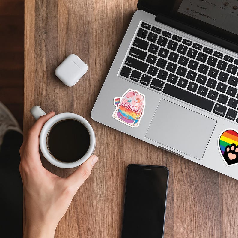 Może przedstawiać: Laptop z naklejką "LGBTea" i tęczowym wzorem. Na drewnianej powierzchni filiżanka kawy, smartfon i bezprzewodowe słuchawki.