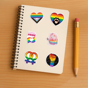 Może przedstawiać: Kolekcja kolorowych naklejek Pride na spiralnym notatniku. Naklejki przedstawiają kształty serc, tęczowe flagi, odciski łap oraz napisy "LGBTIQ+ PRIDE" i "LGBTea". Ż&oacute;łty oł&oacute;wek leży na drewnianej powierzchni.