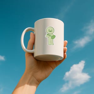 May include: A white ceramic mug held against a blue sky background. The mug features a light green sticker of a cartoon character with a mustache, holding a paintbrush and palette. The mug has a curved handle and a smooth, glossy finish.