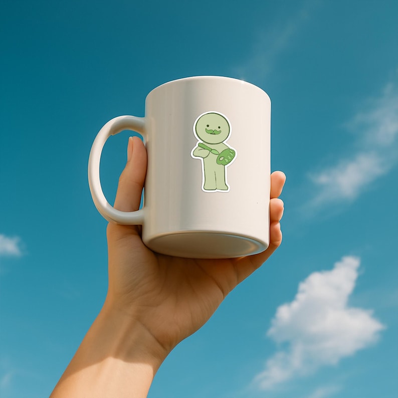 May include: A white ceramic mug held against a blue sky background. The mug features a light green sticker of a cartoon character with a mustache, holding a paintbrush and palette. The mug has a curved handle and a smooth, glossy finish.