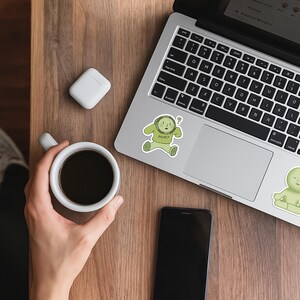 May include: A laptop, smartphone, and a white coffee cup with black coffee are on a wooden surface. A white wireless earbud case and two stickers with a green cartoon character are also visible. One sticker says "HUH?"