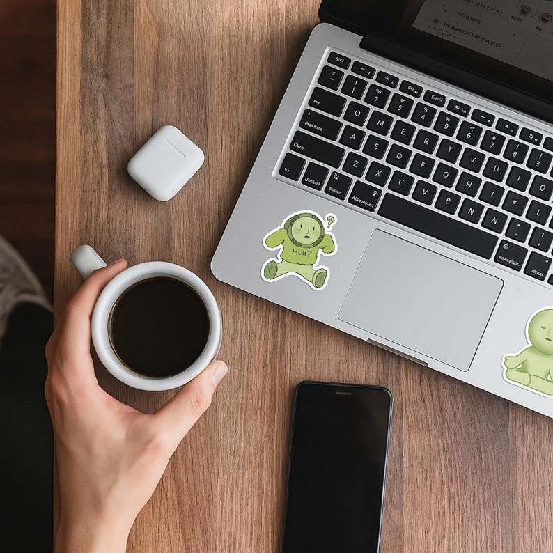 May include: A laptop, smartphone, and a white coffee cup with black coffee are on a wooden surface. A white wireless earbud case and two stickers with a green cartoon character are also visible. One sticker says "HUH?"