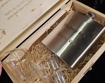 May include: A wooden box with a hinged lid, containing a silver flask engraved with the text "Najbolji kum Dario Vuk" and two clear glass shot glasses. The box is filled with shredded wood.