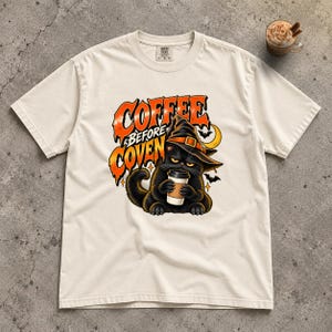 May include: An off-white short-sleeve t-shirt featuring a graphic of a black cat wearing a witch hat and holding a coffee cup. The text reads COFFEE BEFORE COVEN in orange and black lettering, accompanied by small bats and a crescent moon.
