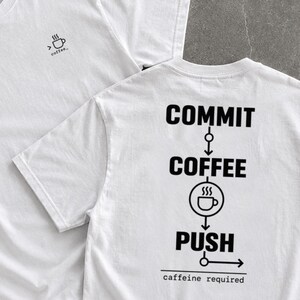 May include: A white t-shirt featuring a small coffee cup icon on the front chest. The back displays a black graphic with the text COMMIT, COFFEE, and PUSH connected by arrows, a circular coffee cup icon, and the text CAFFEINE REQUIRED at the bottom.