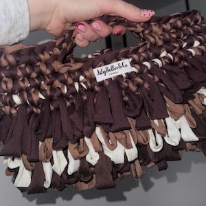 May include: A handmade, fringed handbag in shades of brown and white. The bag features a woven handle and a label that reads "Lily Belles & Co Creations". The fringe adds texture and visual interest to the design.