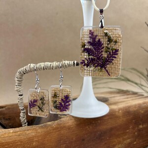 May include: A set of jewelry featuring a necklace and earrings with a rectangular pendant and earrings. The pendant and earrings are made of clear resin with dried purple flowers and a burlap background.