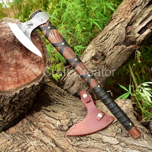 May include: A handcrafted axe with a wooden handle wrapped in black leather and accented with brass studs. The axe head is silver and has a curved blade. A brown leather sheath protects the blade. The axe is displayed on a wooden surface.