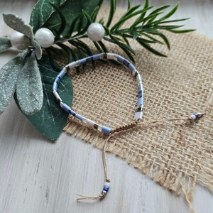 May include: A blue and white beaded bracelet with a brown cord tie. The bracelet is on a burlap surface.
