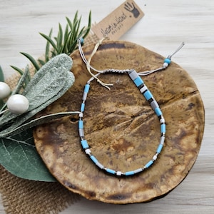 May include: A handmade bracelet with a sliding knot closure. The bracelet features a pattern of blue, gray, white, and silver beads. The bracelet is displayed on a brown, textured surface. A tag with the text "heart in hand designs" is attached.