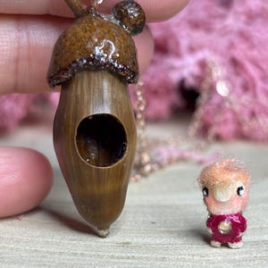 Può includere: Un ciondolo a forma di ghianda marrone con una catena dorata. La ghianda ha un piccolo foro sul lato e un tappo in cima. Il ciondolo è accanto a una piccola statuetta rosa e rossa.