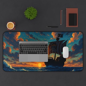 May include: A rectangular desk mat with a pirate ship illustration sailing on a sunset ocean. The ship's sail has a skull and crossbones design. A laptop, mouse, and other desk accessories are on a dark surface, creating a workspace.