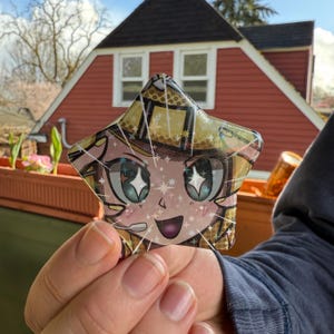 May include: A star-shaped decorative item featuring a cartoon character with large, sparkling eyes and a smiling expression. The design incorporates shades of pink, yellow, and teal, with a glittery effect. The item is held up against a blurred outdoor background.