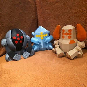 Può includere: Tre figure geometriche colorate sono su una superficie marrone. Le figure sono un robot grigio e nero con accenti rossi, una figura blu a forma di cristallo con accenti gialli e un robot beige con accenti arancioni.