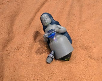 May include: A gray figurine of a woman holding a large blue gemstone. The figurine is sitting on a sandy surface.