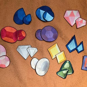 May include: A collection of colorful, embroidered gemstone patches in various shapes and sizes. The patches feature shades of blue, pink, red, purple, yellow, green, and white, with black outlines. The patches are arranged on a textured, orange-brown surface.