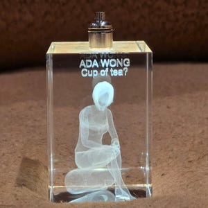 Pode incluir: Um bloco de cristal retangular transparente com uma figura feminina sentada gravada no interior. O texto "ADA WONG Cup of tea?" está acima da figura. Uma tampa prateada está no topo.