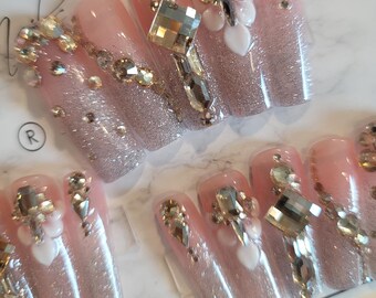 May include: Set of ten false nails with a pink base and glitter. Each nail is decorated with rhinestones and small white hearts.