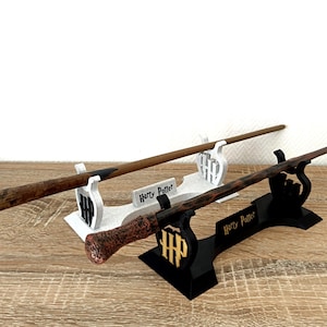 Op de afbeelding: Twee gedetailleerde toverstokken worden op standaards gepresenteerd. Eén toverstok is bruin met een zwarte punt, de andere heeft een rood en bruin handvat. De standaards zijn wit en zwart, met de tekst "Harry Potter".