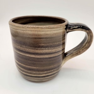 Könnte beinhalten: Eine braune und beige Keramiktasse mit einem gestreiften Muster. Die Tasse hat einen schwarzen Henkel.