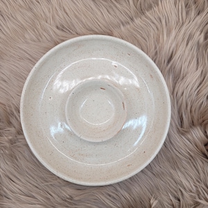 Könnte beinhalten: Eine gesprenkelte, cremefarbene Keramik-Chip- und Dip-Servierschale. Die Schale hat ein kreisförmiges Design mit einer erhöhten Mitte für Dip und einem umlaufenden äußeren Ring für Chips. Die Keramik hat ein rustikales, handgefertigtes Aussehen.
