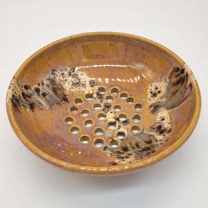 Op de afbeelding: Een handgemaakte keramische zeepbakje met een gespikkelde, geglazuurde afwerking. Het ovale bakje heeft een reeks afvoergaten in het midden. Het kleurenpalet omvat tinten bruin, beige en zwart. Dit functionele stuk is ontworpen voor de badkamer.