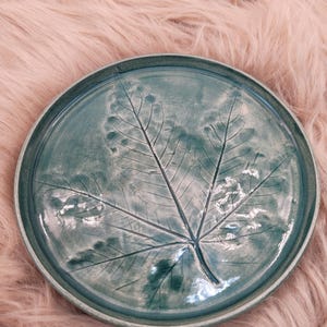 Può includere: Un piatto in ceramica turchese con un'impronta di foglia. Il piatto ha un bordo rialzato e una finitura lucida. Il disegno della foglia è centrato sul piatto e presenta venature dettagliate. È un oggetto decorativo.