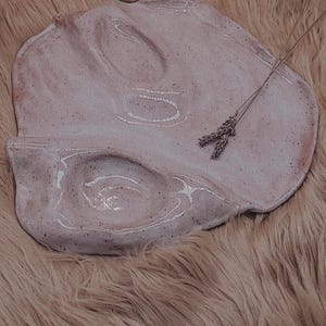 May include: A handmade, organic-shaped ceramic plate in a soft pink hue with speckled detailing. The plate features a unique, undulating design with subtle indentations. A small bundle of dried lavender adds a natural touch.