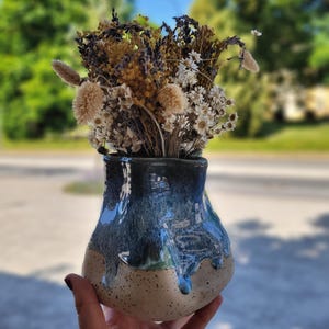 Könnte beinhalten: Eine handgefertigte Keramikvase mit gesprenkeltem beigem Sockel und glänzender, tropfender blauer Glasur. Die Vase enthält einen Strauß getrockneter Blumen in Beige-, Braun- und Weißtönen, wodurch eine natürliche, rustikale Ästhetik entsteht. Die Vase wird in einer Hand gehalten.