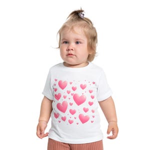 Puede incluir: Una camiseta blanca con un estampado de corazones rosas de diferentes tamaños. Los corazones están dispersos por toda la camiseta, creando un diseño alegre. La camiseta de manga corta parece estar hecha de un material suave.