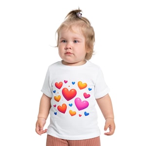 Puede incluir: Una camiseta blanca para niños pequeños con un diseño de corazones coloridos. Los corazones son de color rojo, rosa, naranja, amarillo y azul. La camiseta es de manga corta y tiene cuello redondo. El niño lleva pantalones marrones.