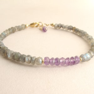 May include: A delicate bracelet made with grey labradorite and amethyst beads. The bracelet has a gold clasp and a small amethyst charm.