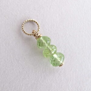 May include: A gold-toned pendant featuring three faceted, light green peridot gemstones. The gemstones are connected and suspended from a twisted gold-filled ring. The pendant is set against a white background.