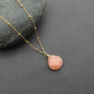 May include: A delicate gold-colored necklace with a peach-colored gemstone pendant. The pendant is teardrop-shaped and wire-wrapped. The chain features small, evenly spaced beads. The necklace is displayed on a dark gray surface.