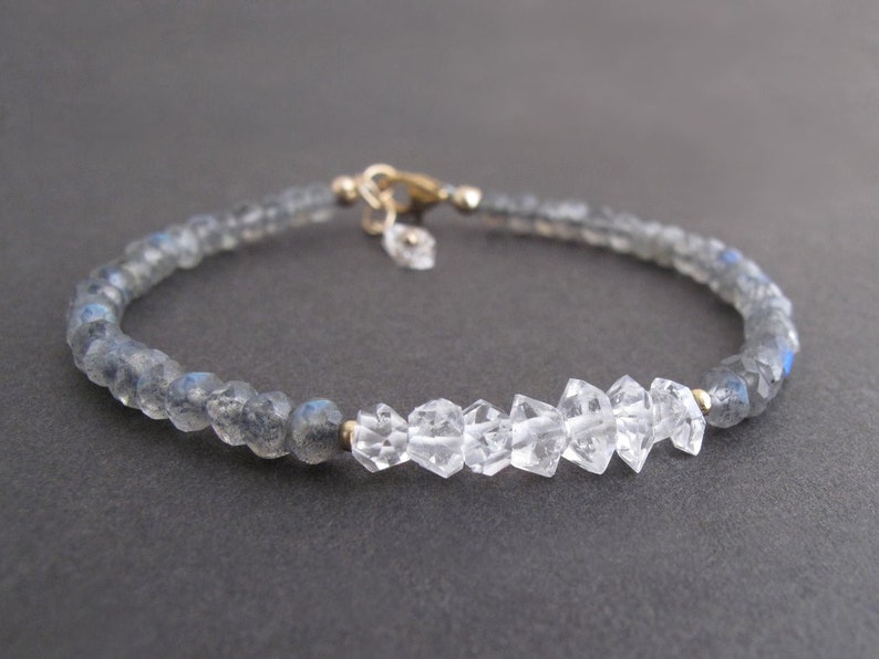 Pu&ograve; includere: Un delicato braccialetto con perline sfaccettate grigio-blu e pietre di cristallo chiare e grezze. Il braccialetto &egrave; assicurato con una chiusura color oro e un piccolo ciondolo, che mostra un design minimalista.