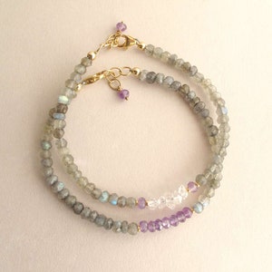 Pu&ograve; includere: Due delicati braccialetti di perline. I braccialetti presentano piccole pietre preziose grigie sfaccettate, intervallate da perline chiare e viola. Chiusure color oro e piccole perline viola completano il design. I braccialetti sono esposti su uno sfondo chiaro.