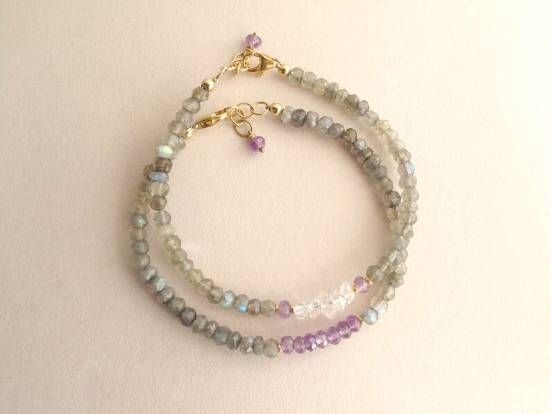 Pu&ograve; includere: Due delicati braccialetti di perline. I braccialetti presentano piccole pietre preziose grigie sfaccettate, intervallate da perline chiare e viola. Chiusure color oro e piccole perline viola completano il design. I braccialetti sono esposti su uno sfondo chiaro.