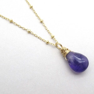 May include: A delicate gold chain necklace with a single purple gemstone pendant. The gemstone is wrapped in gold wire and has a teardrop shape.