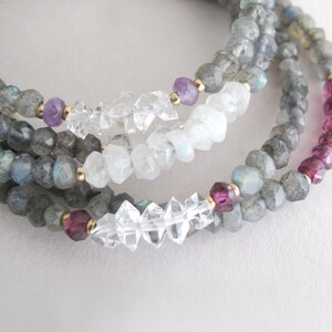 Pu&ograve; includere: Primo piano di collane di pietre preziose a strati. Le collane presentano perline di labradorite grigia, quarzo trasparente, ametista e accenti di tormalina rosa. Perline color oro separano le pietre preziose. Le collane sono esposte su una superficie bianca.