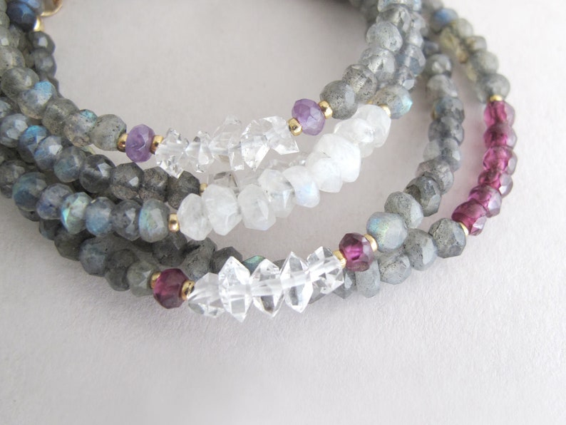 Pu&ograve; includere: Primo piano di collane di pietre preziose a strati. Le collane presentano perline di labradorite grigia, quarzo trasparente, ametista e accenti di tormalina rosa. Perline color oro separano le pietre preziose. Le collane sono esposte su una superficie bianca.